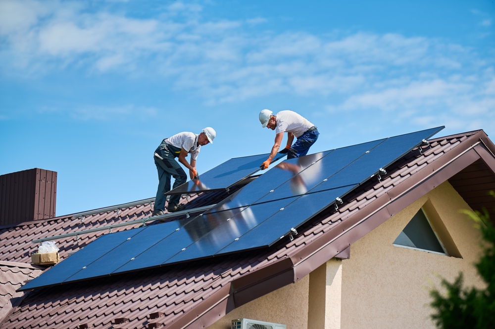 Two workers wearing helmets install solar panels on the roof of a house under a blue sky, showcasing renewable energy efforts.